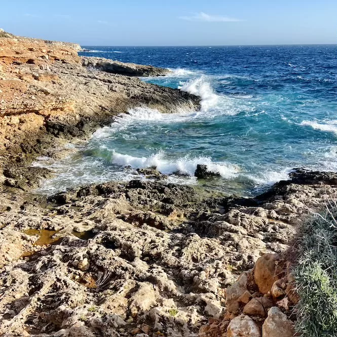 Rocky shoreline in the Hagar Qim Temple Area, with waves crashing against the rugged terrain. The turquoise waters contrast with the weathered rocks, creating a picturesque seascape.