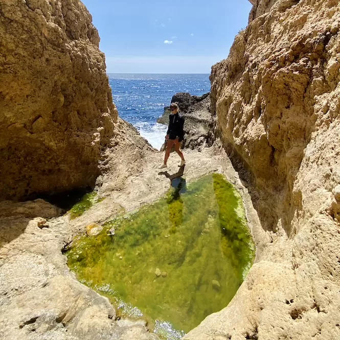 A gorge in Miġra l-Ferħa featuring a secluded rock formations with opening into a sea.  A lone explorer stands amidst the rocky cliffs, enjoying the beauty of this lesser-known spot.