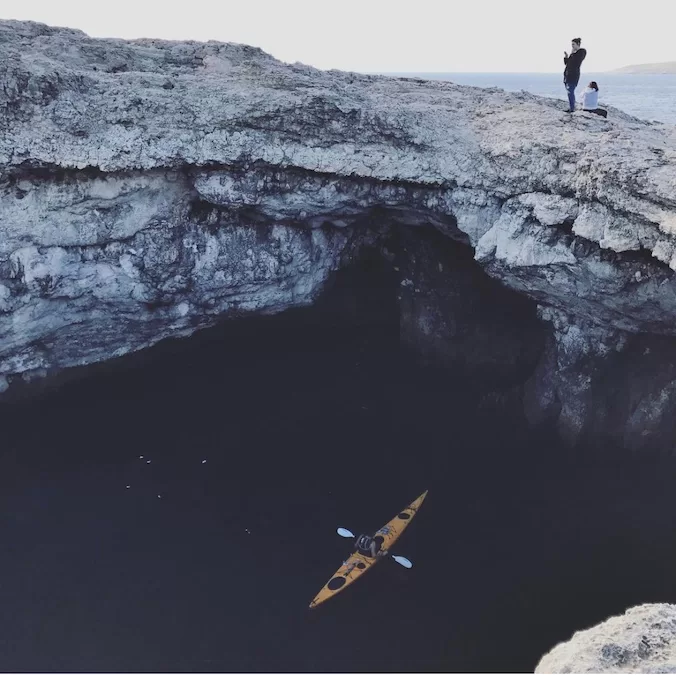 Coral Lagoon, a lesser-known spot in Malta, featuring a natural archway carved into the rocky coastline. A solitary kayak glides over the dark waters.