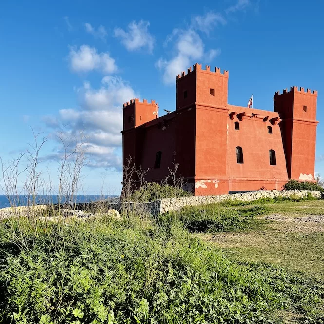 A red fortress-like structure stands prominently against a backdrop of blue sky and scattered clouds. This historical landmark, known as the Red Tower, is located in the North of Malta. It's surrounded by lush greenery.