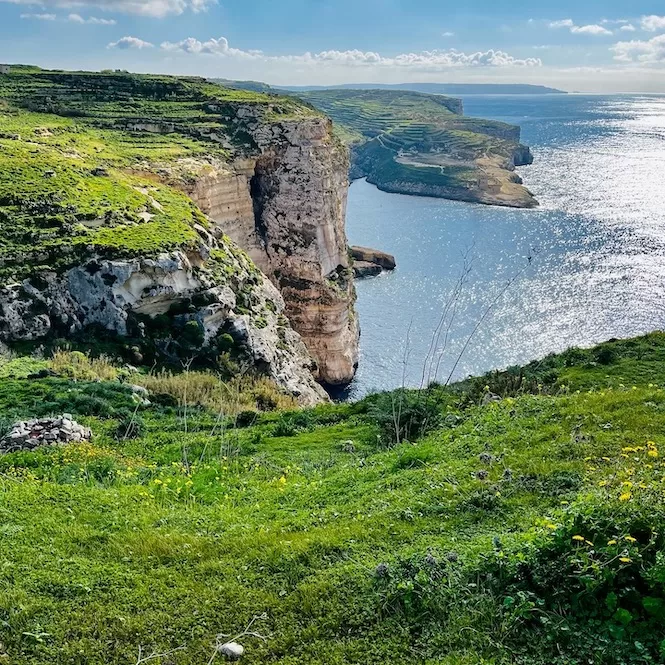 A scenic view of lush green cliffs overlooking the sparkling blue sea, with sunlight reflecting off the water. Don't skip such a hidden gem when in Malta.