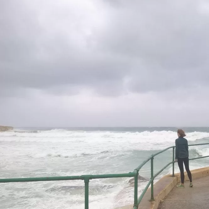 A person standing by a railing, looking out at rough waves crashing against the shore under cloudy skies on a rainy day.