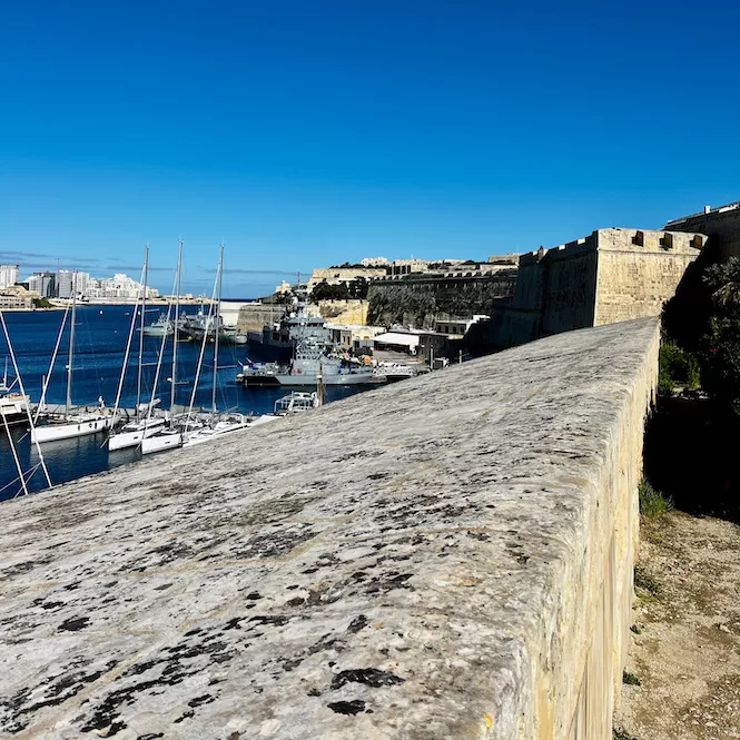 A view from a limestone fortification at Sa Maison Garden overlooking a harbour with sailboats and naval ships.