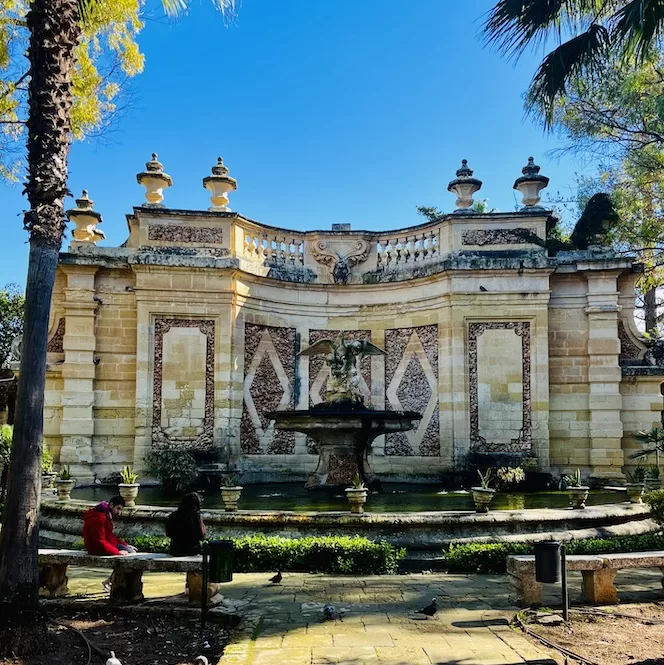 A decorative fountain surrounded by benches and trees in San Anton Gardens on a sunny day.