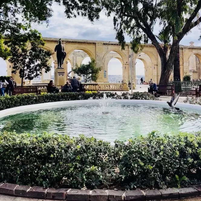 A fountain surrounded by bushes and people sitting on benches in Upper Barrakka Gardens with arched structures in the background.