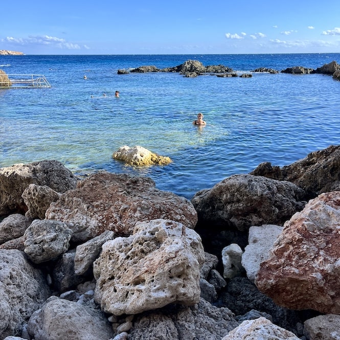 People swimming in the clear, turquoise waters of a rocky Ghar Lapsi Beach. Large boulders dot the shallows, while cliffs are visible in the distance.