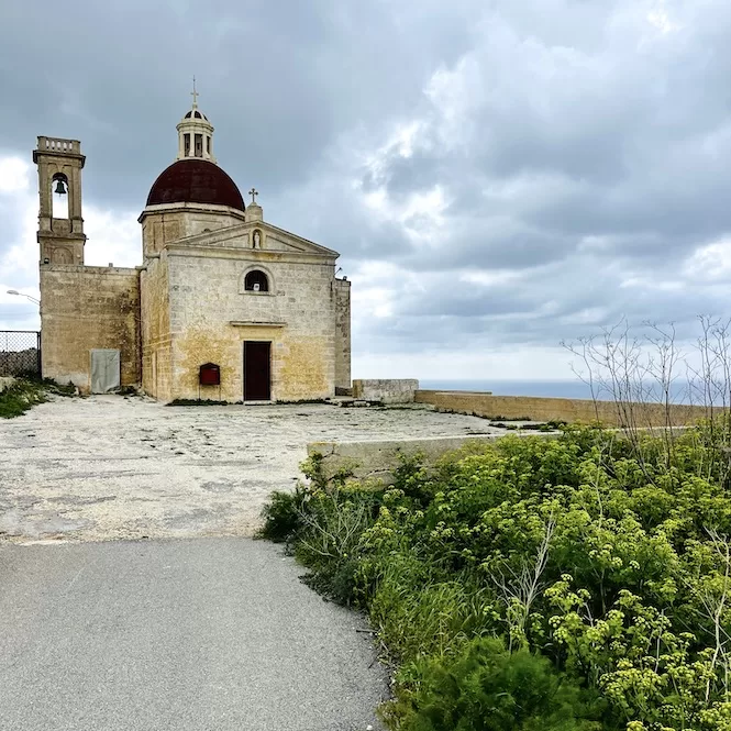 The image shows a Mtahleb Chapel with a red-domed roof surrounded by green bushes under a cloudy sky.