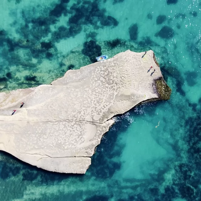 Aerial view of Ta' Babu Cove, isolated rock formation surrounded by the clear turquoise waters of a calm sea. The rock's textured surface contrasts with the sea's natural patterns, creating a tranquil escape for a small group enjoying the sun.