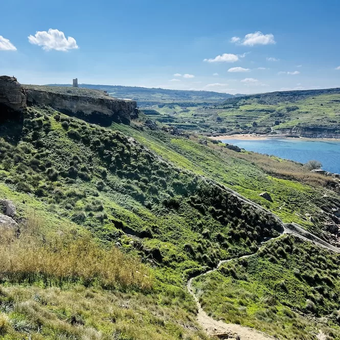 A scenic landscape on a hike from Għajn Tuffieħa Beach to Gnejna Bay with rolling green hills, rocky cliffs, and a sea in the distance. A narrow path winds through the grassy hillside in the foreground. In the background, there is a limestone watchtower on top of a cliff overlooking the bay.