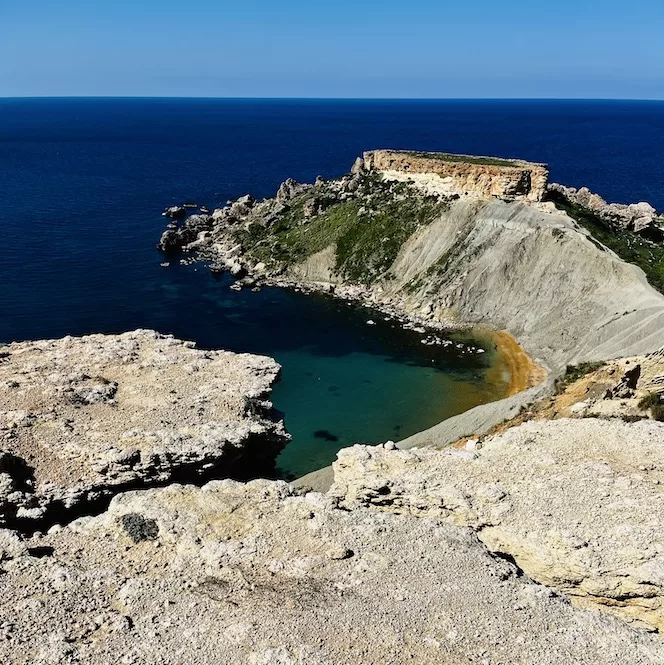 The image shows a rugged and dramatic coastal landscape. In the foreground, there are jagged rocky cliffs and formations surrounding a small, emerald green il-Qarraba Peninsula. The cliffs rise up sharply with patches of vegetation growing on them. In the background is the deep blue of the open sea stretching out to the horizon under a clear blue sky.
