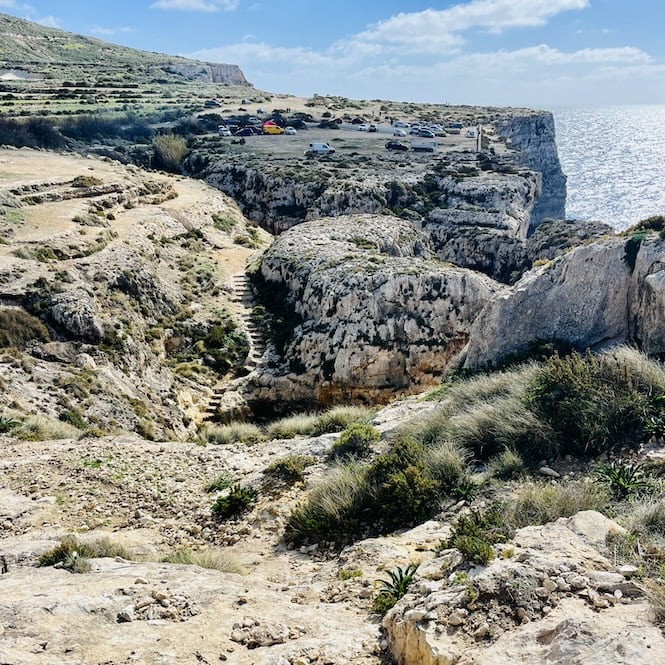 A rugged coastal landscape with rocky cliffs and sparse vegetation of Miġra l-Ferħa area. A narrow path winds its way down the cliffside, leading to a rocky shoreline. Several cars are parked on the cliff edge above.