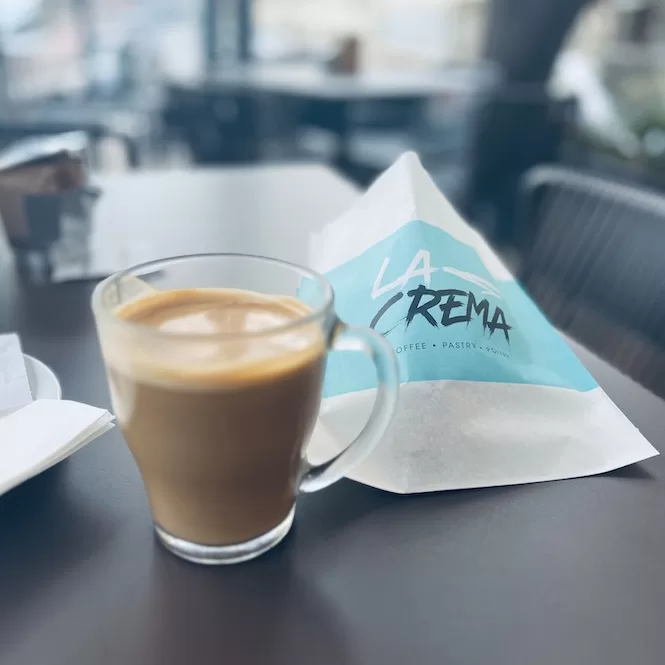 A cup of coffee and a pastry bag from La Crema in Sliema. The table setting suggests a cosy cafe ambience, inviting customers to enjoy a delightful coffee break.