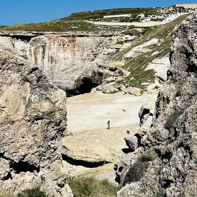 This image shows Il-Blata tal-Melh, a rugged landscape with rocky cliffs and a dry limestone plateau. Two people are visible in the distance, highlighting the scale of the terrain.