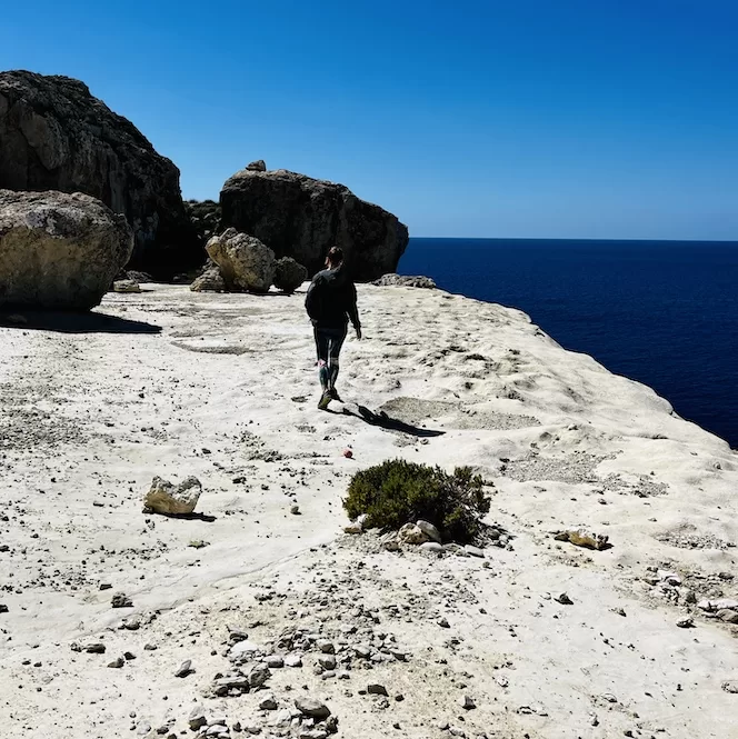 A person is walking along the rocky cliffs of Il-Blata tal-Melh, with the vast blue ocean stretching out in the distance.