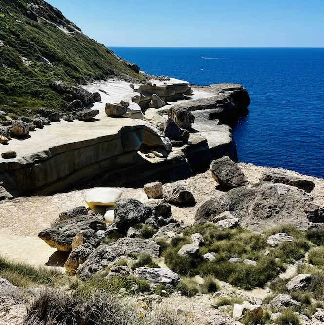 A rocky landscape of Il-Blata tal-Melh in front of the vast expanse of the ocean. The rugged terrain is lined with large boulders and patches of greenery, creating a striking contrast against the deep blue sea.