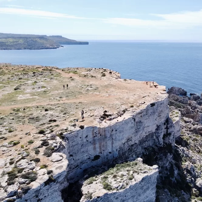 A vast expanse of rugged cliffs overlooking the sea at Majjistral Park. Several people are seen exploring the rocky terrain, surrounded by the blue waters of the sea.