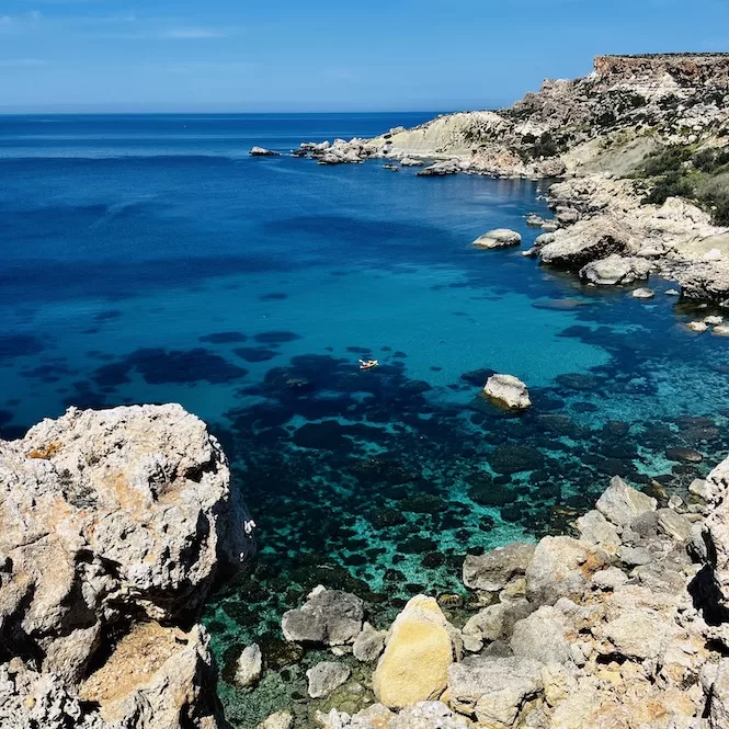 A serene view of the crystal-clear waters of Majjistral Park, with rocks scattered beneath the surface and a lone kayak enjoying the sea.