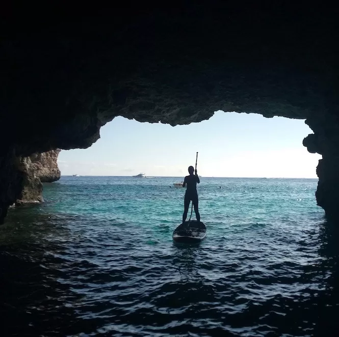 A person standing on a paddle board inside a cave, with sunlight illuminating the turquoise waters outside and boats visible in the distance.