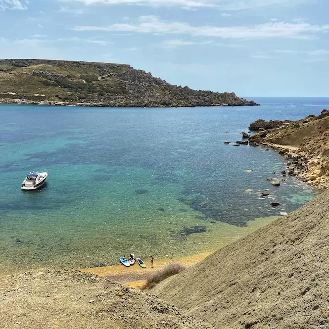 Exploring the coast from Ghajn Tuffieha to Gnejna Bay, showing a serene coastal scene with clear turquoise waters, a small motorboat, and people standing near paddle boards on the sandy shore.






