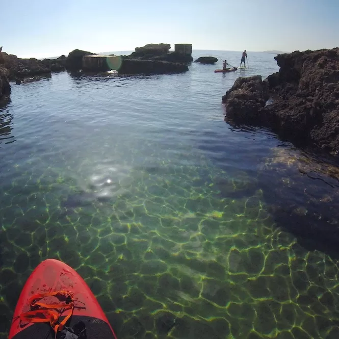 A red kayak in the foreground with crystal-clear waters revealing a person paddle boarding in the distance among rocky formations at Slugs Bay, Malta.
