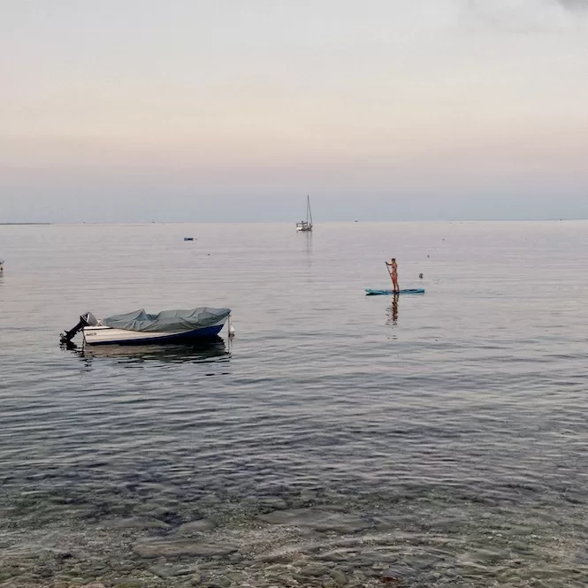 A person standing on a paddle board in calm waters of St. Thomas Bay as the sun sets, with boats in the background.