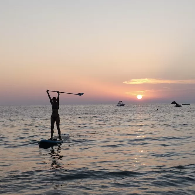 Capturing the silhouette of a person standing on a paddle board in Golden Bay, Malta against the backdrop of the setting sun and calm sea waters, with boats visible in the distance.