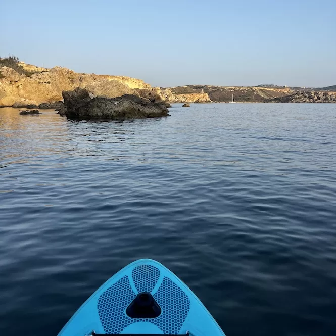 An image taken by a paddle boarder sitting on their board in calm, crystal-clear blue water. A rugged coastline and rocky cliffs Golden Bay are visible in the background under a clear sky.