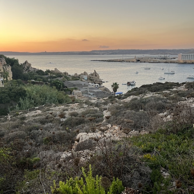 A landscape overlooking a rocky coastline with sparse vegetation, boats anchored in the calm waters, and the setting sun casting a warm glow over the scene at Paradise Beach.