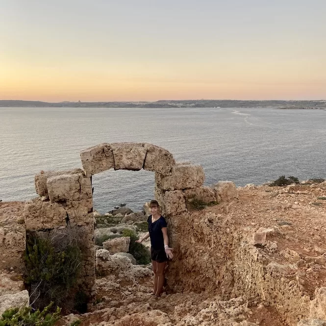 A person standing next to a limestone window on a rocky cliff overlooking the calm sea during sunset.