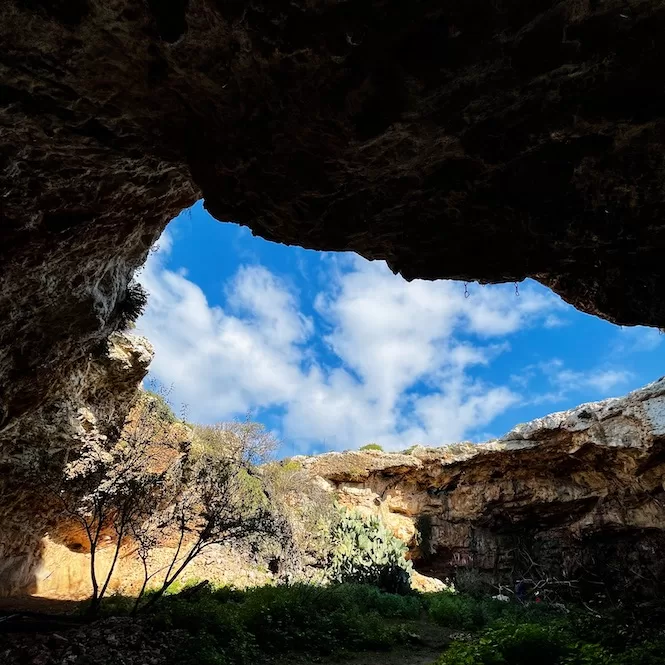 A picturesque view from within a cave, showcasing the blue sky adorned with fluffy clouds, surrounded by the rocky landscape and lush greenery.