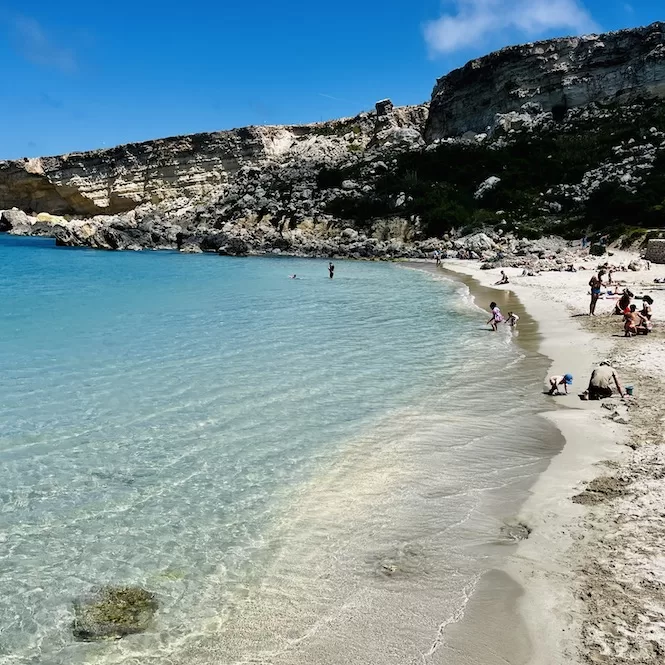 Families enjoy the sunny day at the Paradise Beach, with some playing in the shallow waters and others relaxing on the sandy shore.