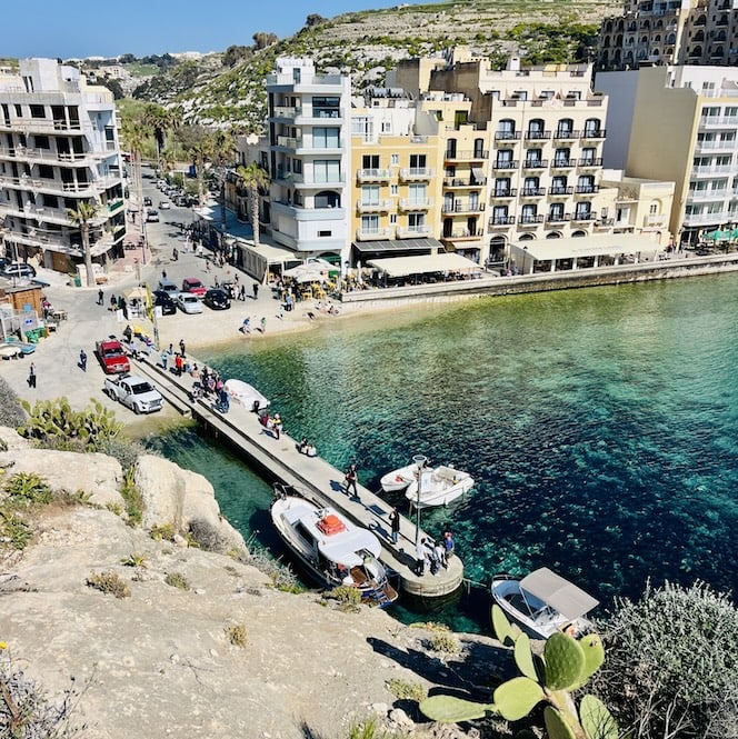 Overlooking Xlendi, with buildings lining the shore and boats docked along the pier. The crystal-clear turquoise water contrasts against the sandy beach, while people stroll along the waterfront.