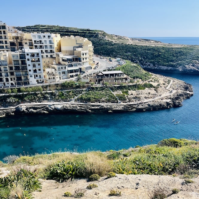 A panoramic view of Xlendi Bay seen from the top of the cliffs. The bay is surrounded by buildings and lush greenery, with crystal-clear turquoise water stretching out to the horizon.