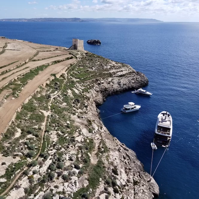 Aerial view of Mgarr Ix-Xini Tower overlooking the scenic coastline of Gozo, Malta. Two yachts are anchored in the blue waters near the rocky shore.