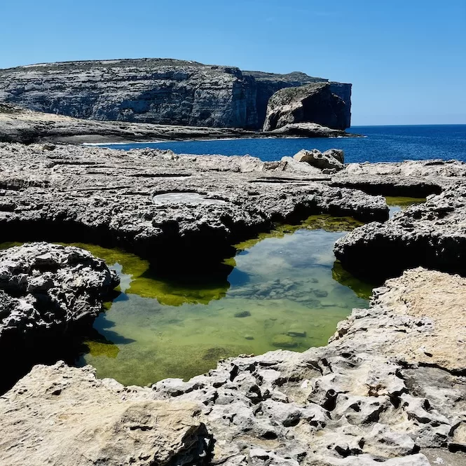 Rocky pools dotting the landscape offer the view of Dwejra Bay in Gozo. Beyond, the majestic Fungus Rock stands tall against the vast blue expanse of the sea.