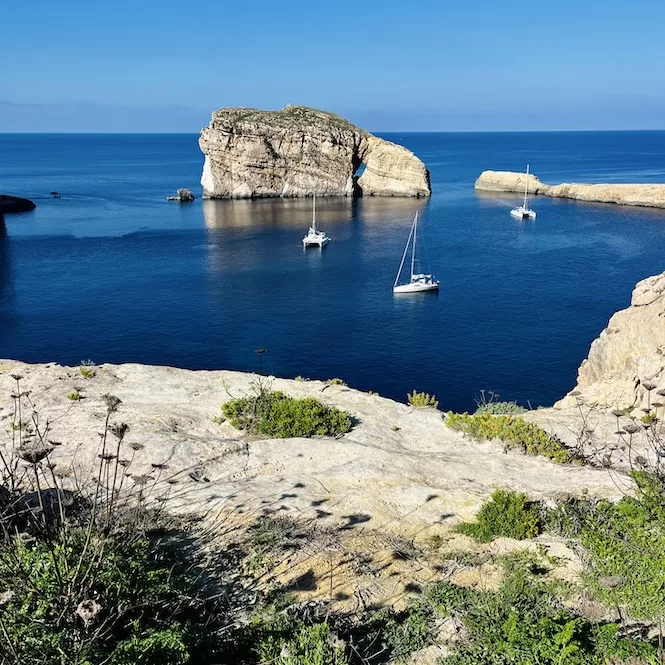 Sailboats are anchored in the calm waters of Dwejra Bay, near the Inland Sea, Gozo, with rocky cliffs in the background.