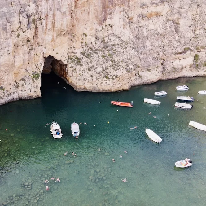 Aerial view of the Tunnel of the Inland Sea in Gozo, with boats floating on the clear turquoise water and people swimming. 