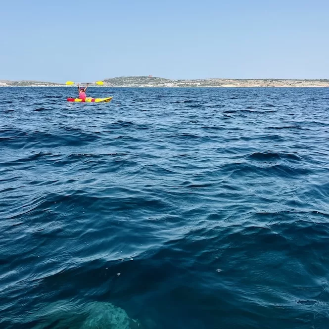 Kayaking in Ghadira Bay, the clear blue waters stretch endlessly, with the adventurer in the red kayak enjoying the vast expanse of the sea under the bright sun.