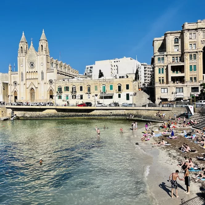 People enjoying a sunny day at Balluta Bay in St. Julian's, Malta, with swimmers and sunbathers by the beach and a prominent church in the background.