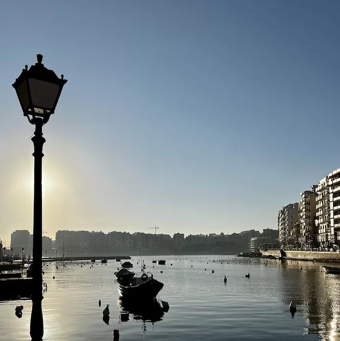 A serene morning scene at St. Julian's Bay, with boats peacefully floating on calm waters against a backdrop of buildings and a silhouette of a street lamp.