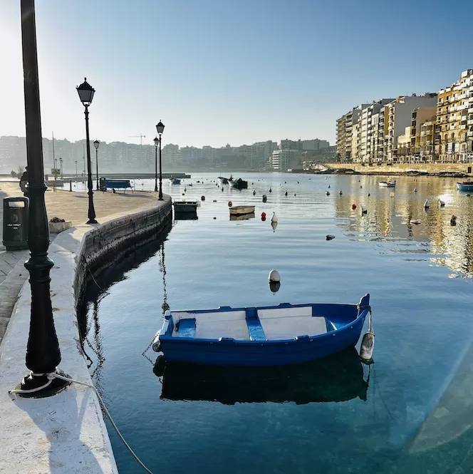 A blue boat anchored in calm waters at Spinola Bay, St. Julians, Malta, with buildings lining the waterfront in the background.