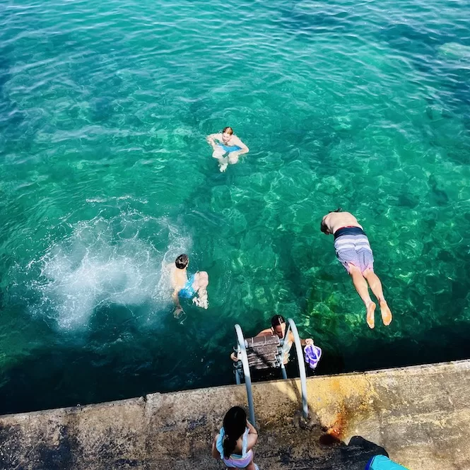 People swimming in the crystal-clear waters of St. Julians, Malta, with one person diving off a ladder into the sea.