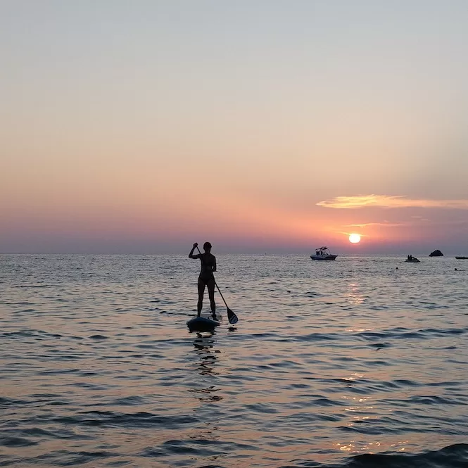 Silhouetted figure paddle boarding on the calm waters of Ghajn Tuffieha beach in Malta as the vibrant hues of the sunset paint the sky in shades of orange and pink.