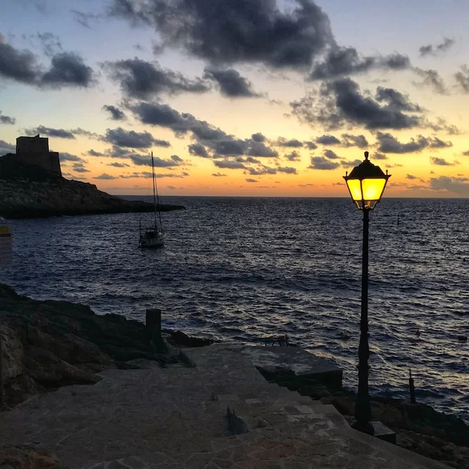 A lamppost illuminating the evening sunset scene at Xlendi, Gozo, as the sun sets over the horizon, casting a warm glow across the sky and reflecting gently on the waters, with a sailboat peacefully anchored in the distance.