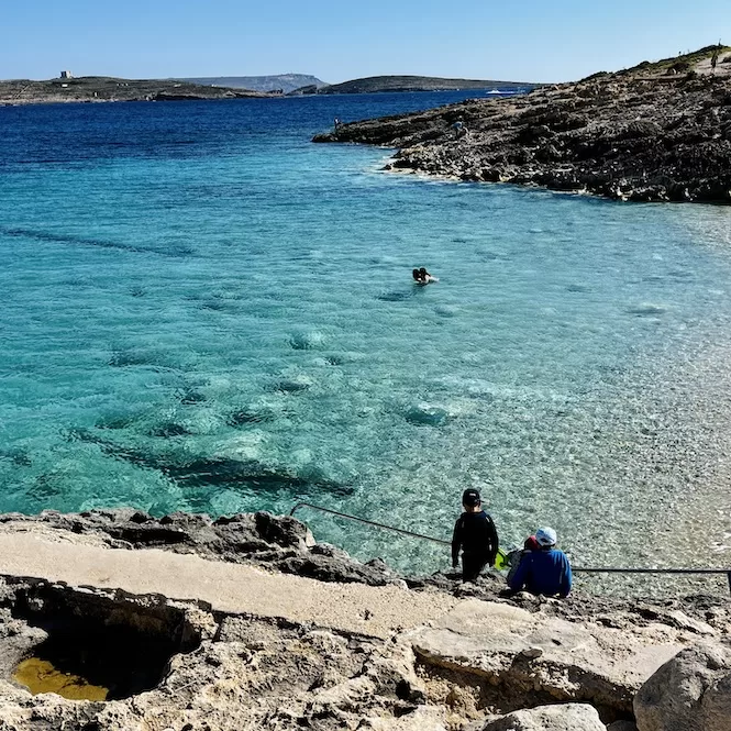 People sitting on rocky steps overlooking the crystal-clear waters of Hondoq ir-Rummien Bay in Gozo, Malta, with a swimmer enjoying the refreshing sea.