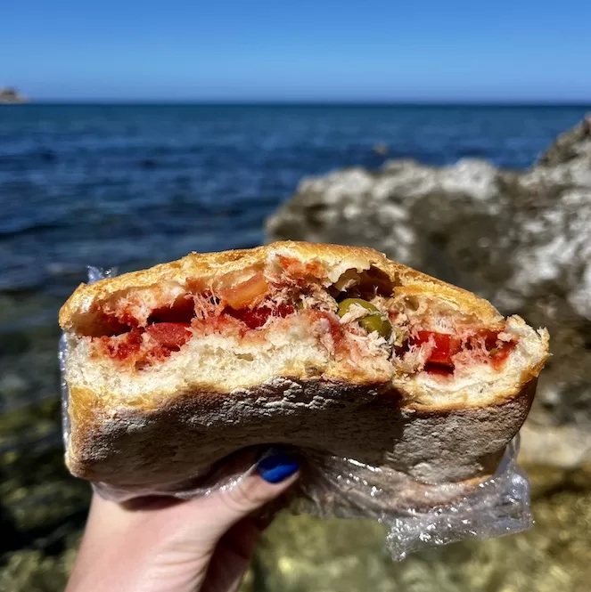 A half-eaten ftira biż-żejt sandwich held against a backdrop of rocky shorelines and ocean waves. Ftira biż-żejt is a traditional Maltese snack made with bread filled with tomatoes, tuna, olives, and capers. It's a widely available and affordable food found throughout Malta and Gozo.