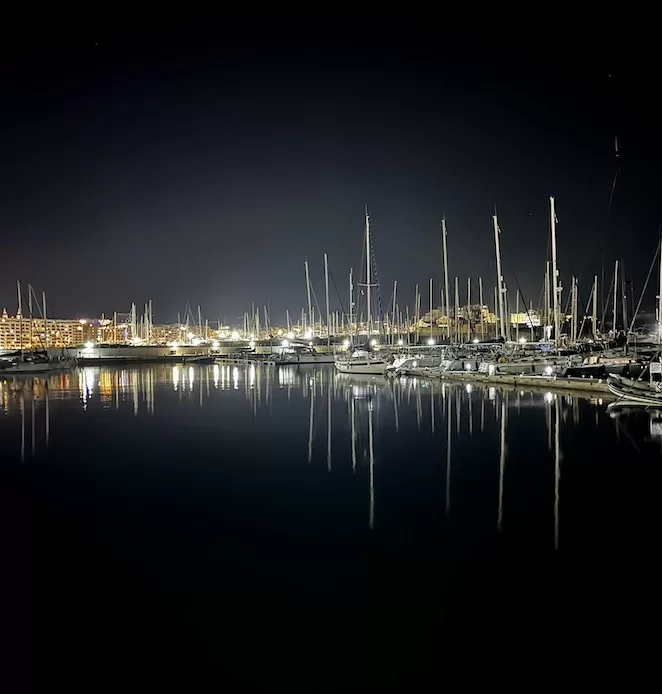 A night view of a Gzira marina filled with sailboats, their reflections shimmering on the calm water. The lights from the boats and surrounding buildings create a atmosphere, perfect for a leisurely night walk