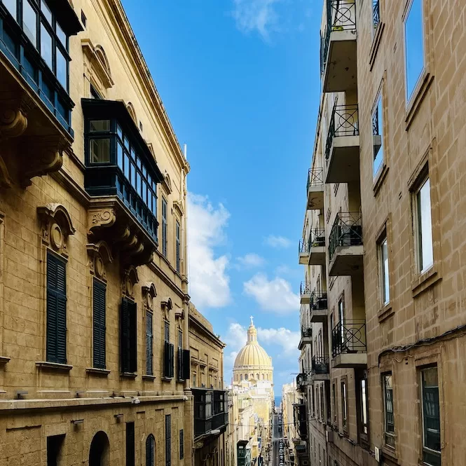 A narrow street lined with traditional Maltese buildings in Valletta, with the dome of a historic church visible at the end of the road under a blue sky with clouds.