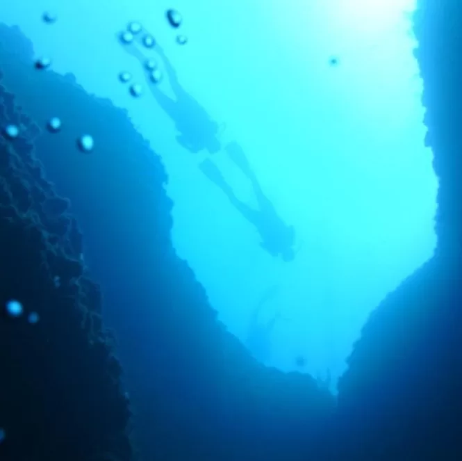 Two scuba divers are submerged in deep blue water, descending into a rocky underwater landscape. Sunlight filters through the water, casting a soft glow around them. Bubbles rise from their breathing apparatus as they explore the underwater environment. This image captures the adventure of scuba diving, a popular water sport in Malta.
