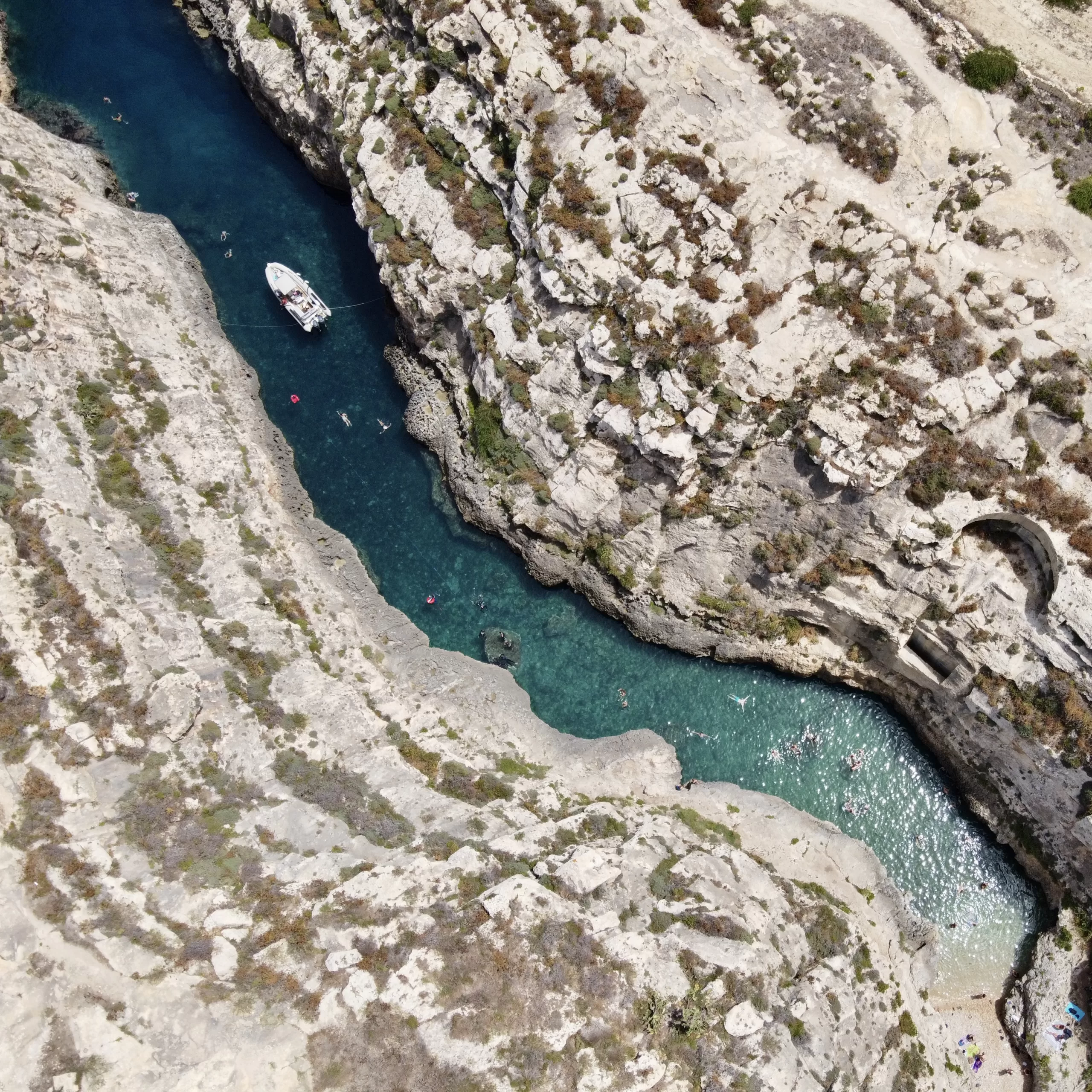 Aerial view of a rocky coastline with turquoise waters of Ghasri Valley in Gozo, Malta. A boat is anchored near the cliffs, and people are snorkelling in the clear water.